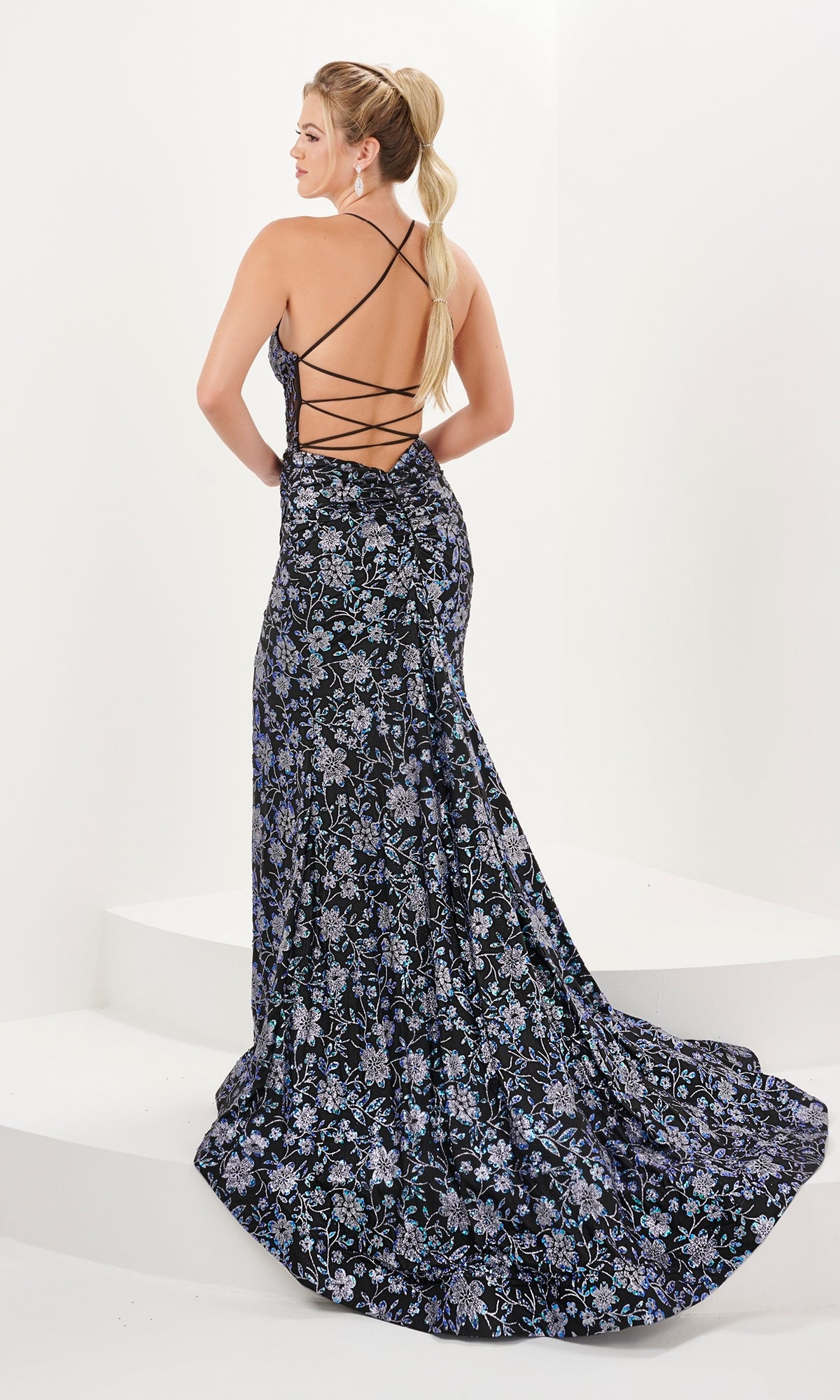 A woman in the Long Prom Dress 16060 by Tiffany—floor-length with a floral print, low back, and crisscross straps—stands on white steps, facing away from the camera.